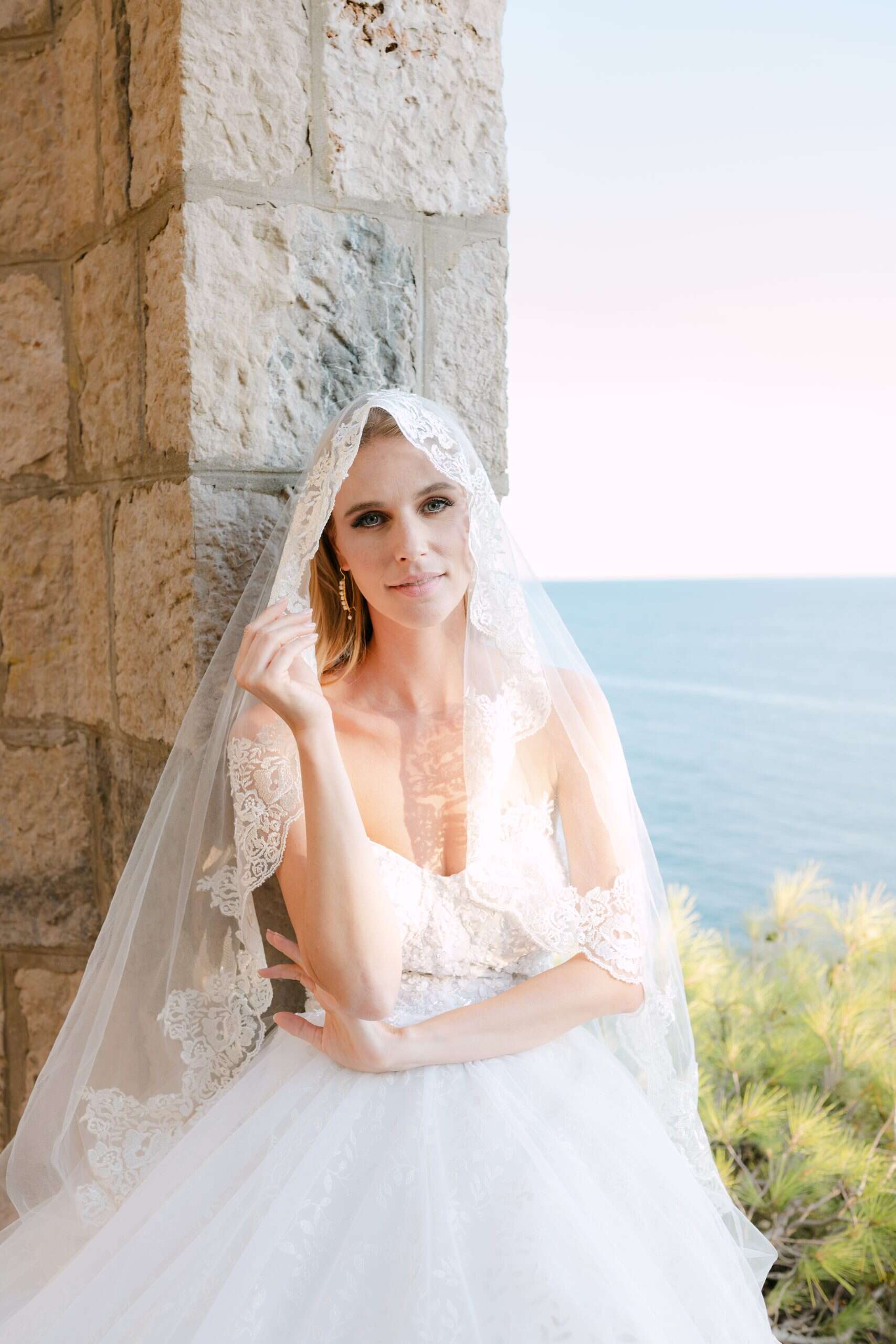 Prekrasna mladenka pozira naslonjena na kameni zid s raskošnim čipkastim velom prebačenim preko glave. Opušteni ljetni destination wedding portret obasjan suncem.