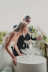 Mladenci se zabavljaju uz kulu od čaša za šampanjac. Mladenka miriše šampanjac iz čaše na vrhu kule dok mladoženja drži bocu tijekom proslave vjenčanja u Dubrovniku.