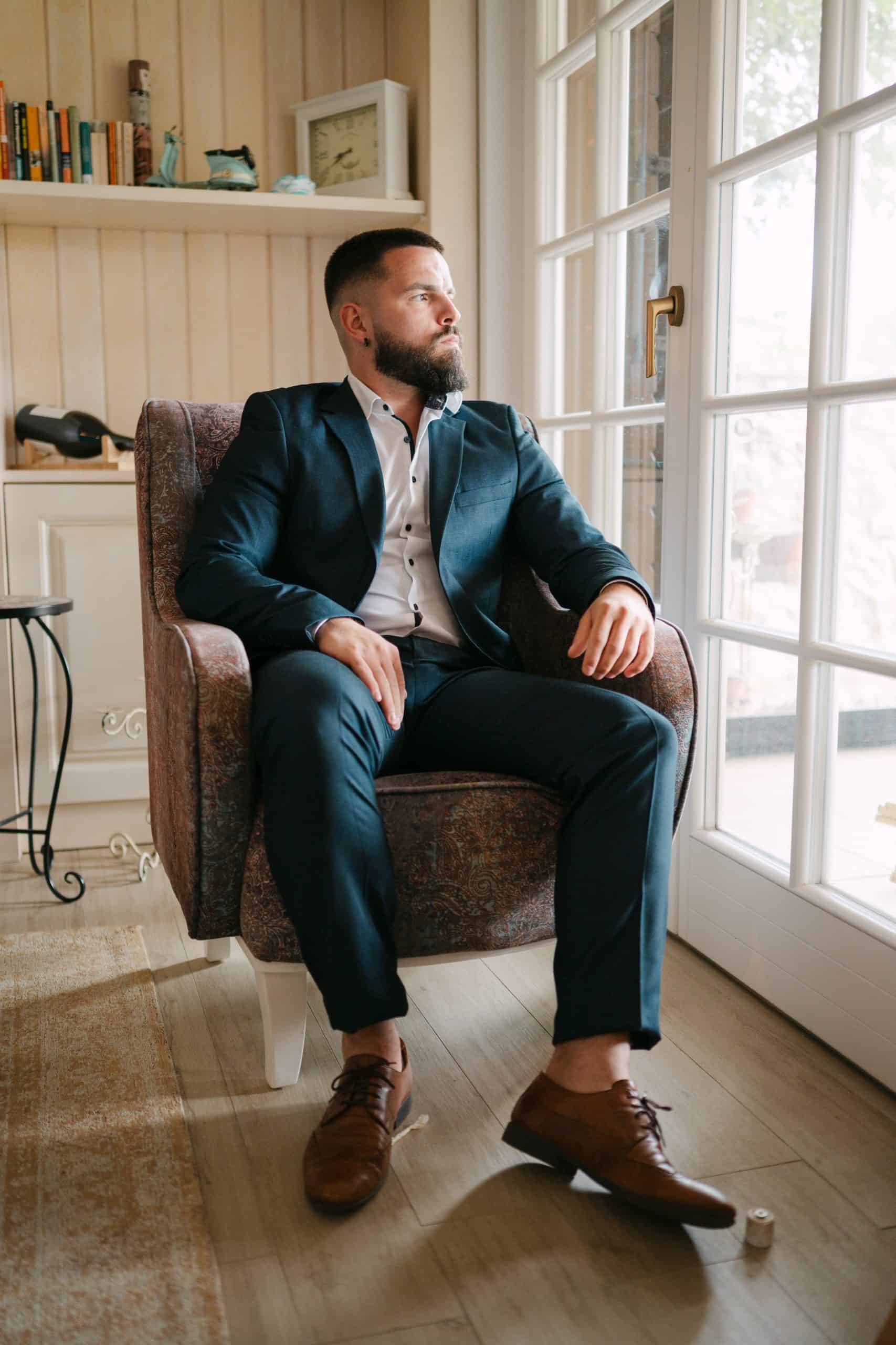 Elegantan portret mladoženje u tamnom odijelu koji sjedi u fotelji pored prozora. Profinjena fotografija priprema za vjenčanje.