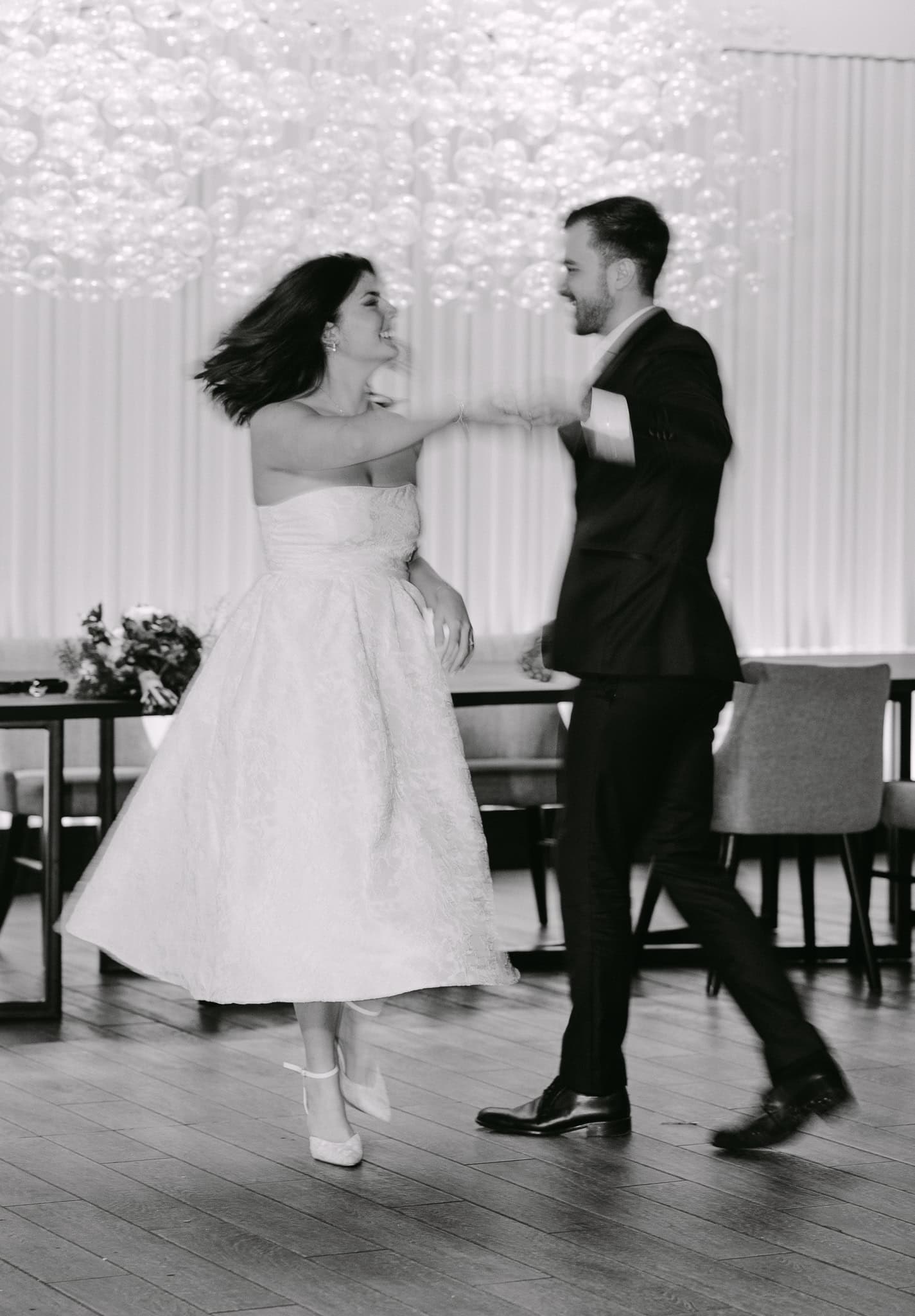 Mladenci plešu svoj prvi ples u elegantnom ambijentu. Crno-bijela fotografija s fokusom na pokret i radost mladenaca.