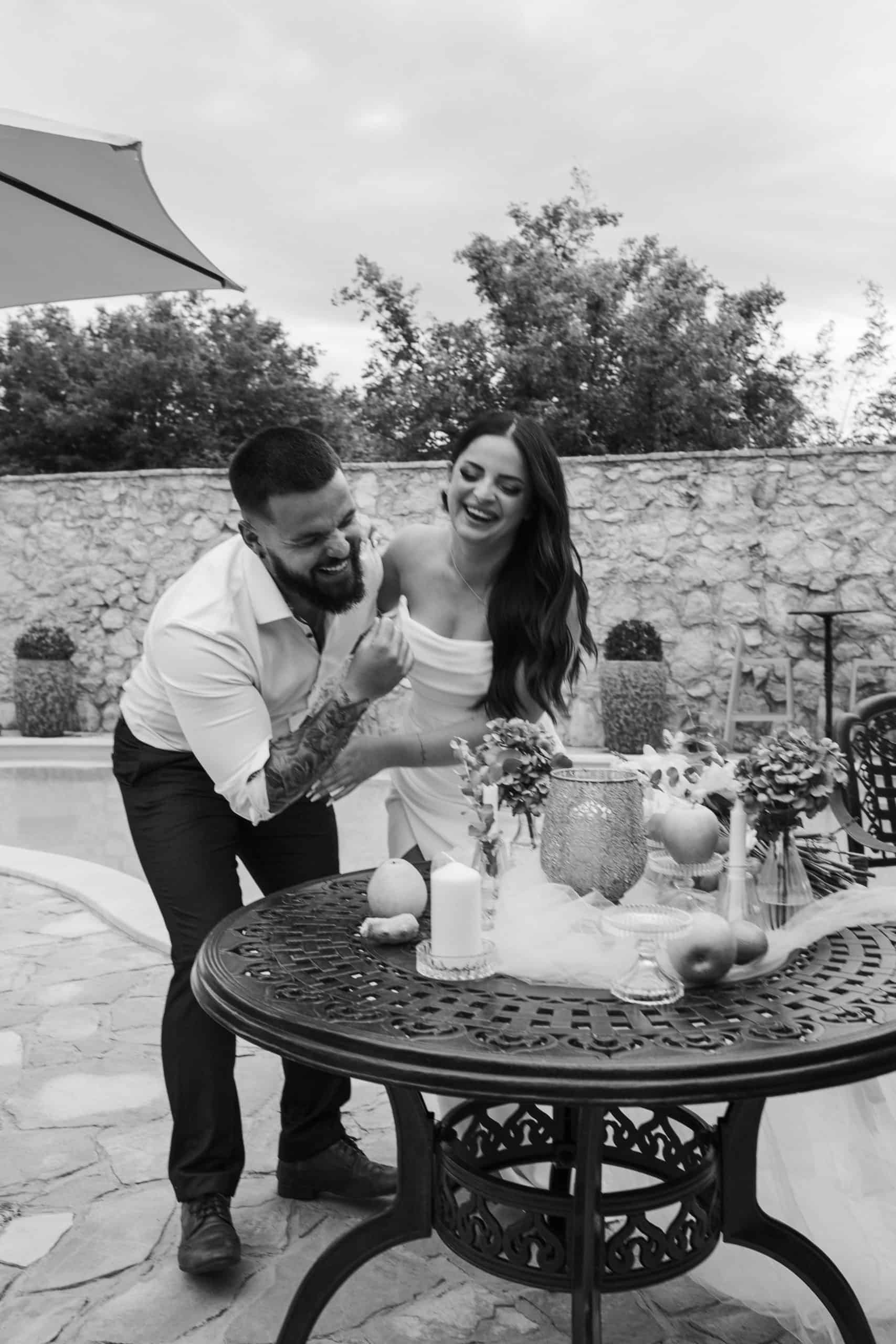 Crno-bijela fotografija mladenaca koji se spontano smiju za stolom pored bazena. Dokumentarni stil vjenčane fotografije pun emocija.