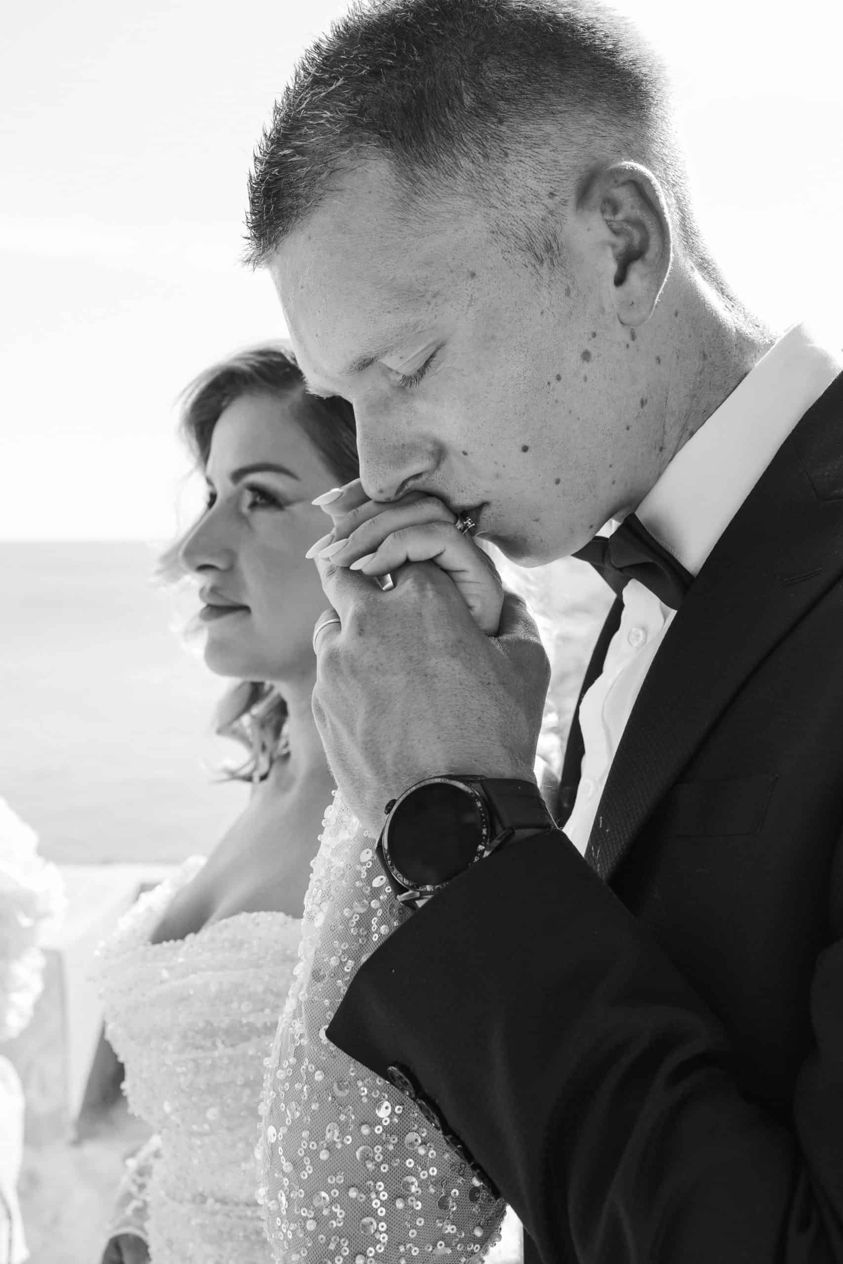 Crno-bijeli krupni kadar mladoženje koji nježno ljubi ruku mladenke. Oboje gledaju u daljinu na destination vjenčanju obasjanom ljetnim suncem.