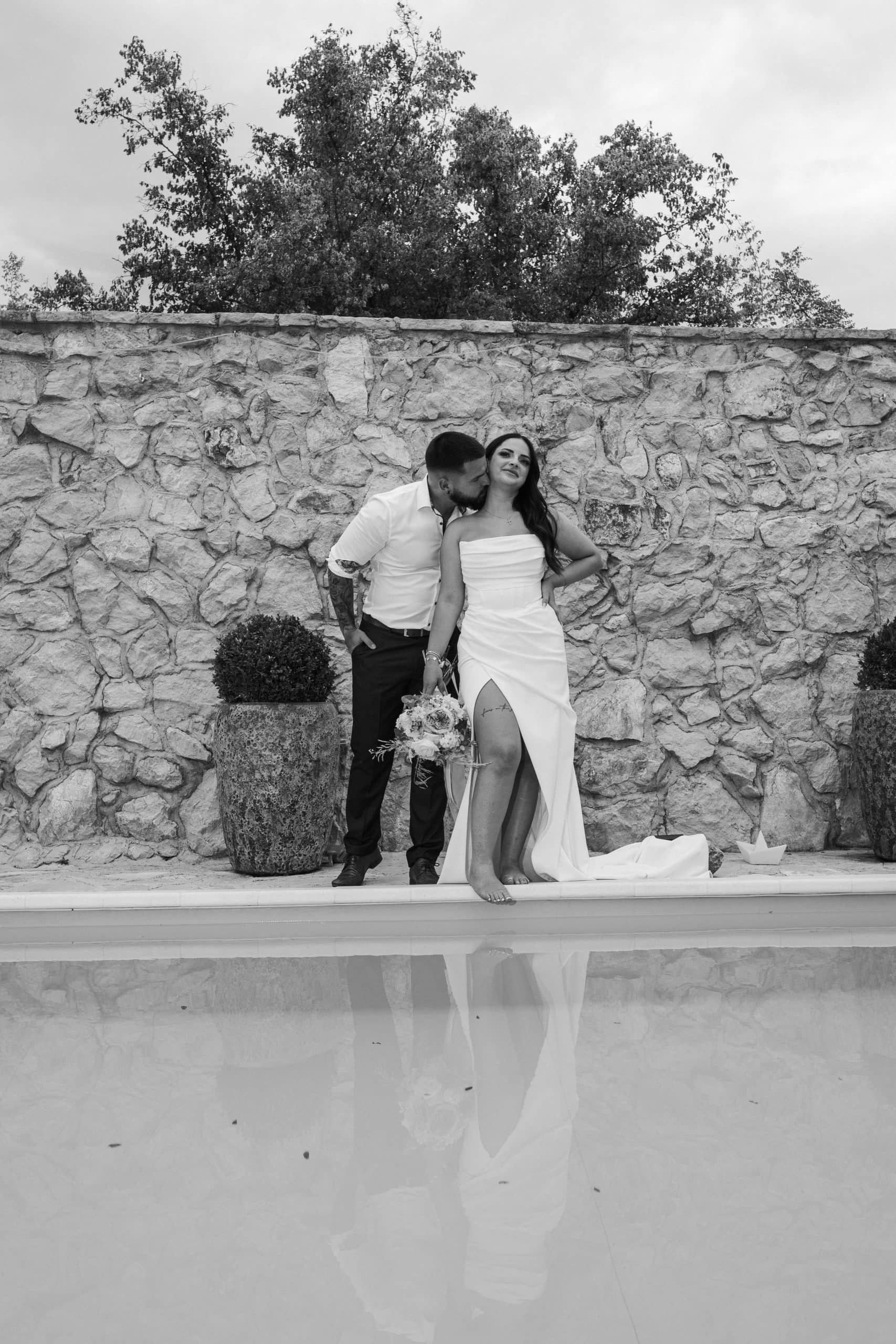 Mladenci poziraju ispred rustikalnog kamenog zida pored bazena. Umjetnička crno-bijela fotografija vjenčanja s odrazom u vodi.