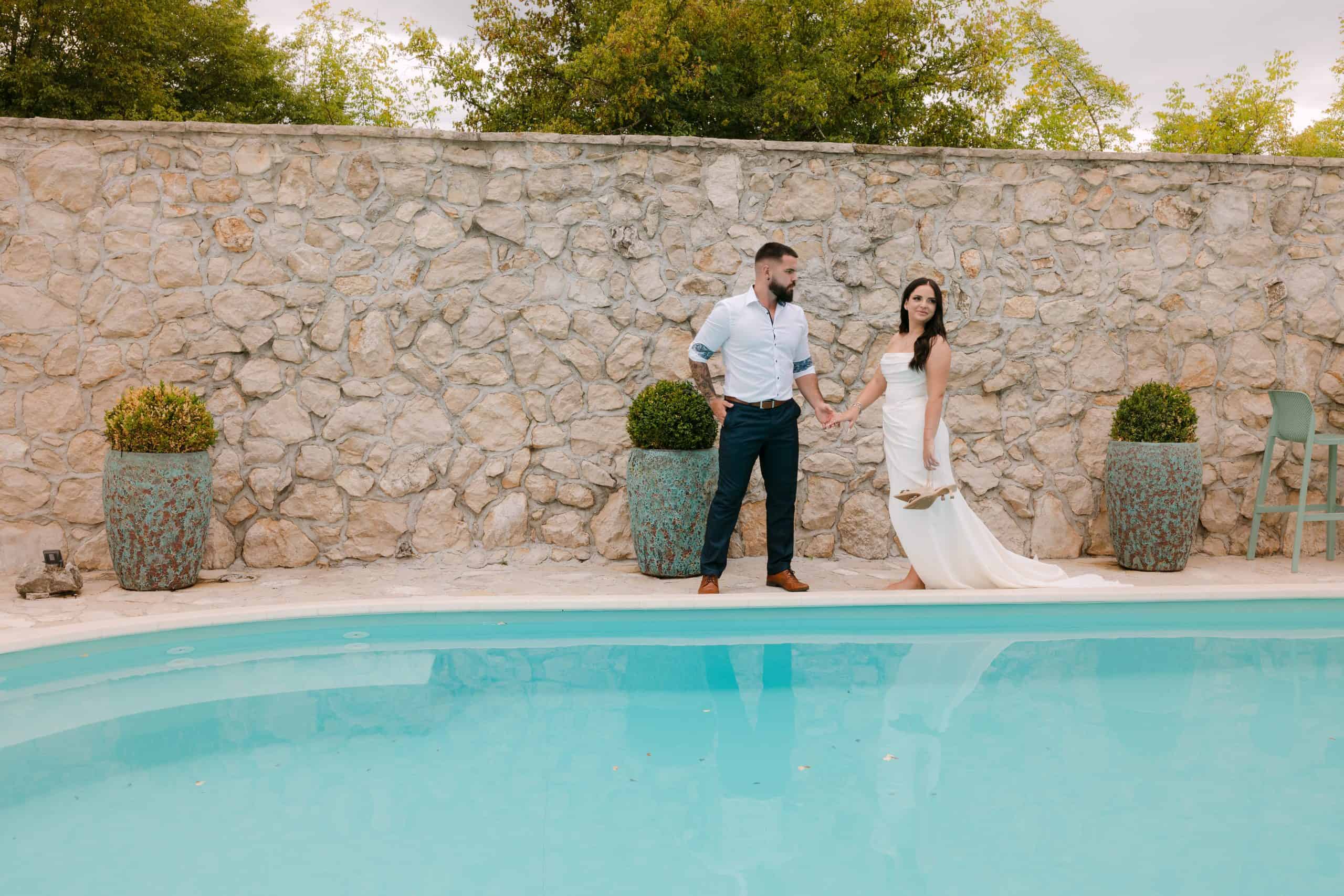 Mladenci poziraju pored bazena ispred kamenog zida. Moderna i minimalistička vjenčana fotografija u mediteranskom stilu.