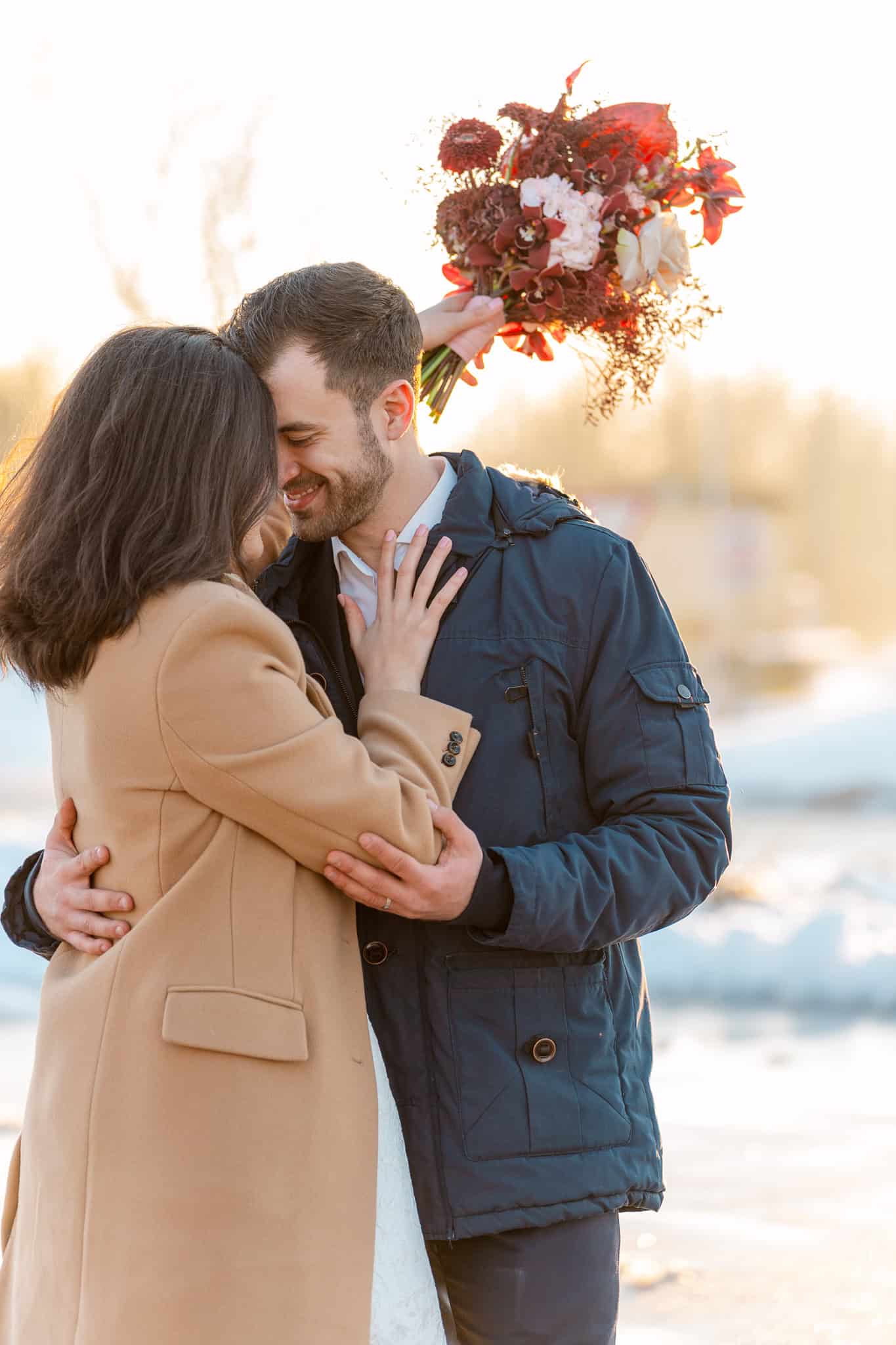 Mladenci u toplim kaputima u nježnom zagrljaju tijekom zimskog sessiona. Mladenka drži bogati crveni buket cvijeća dok ih obasjava toplo zimsko sunce.