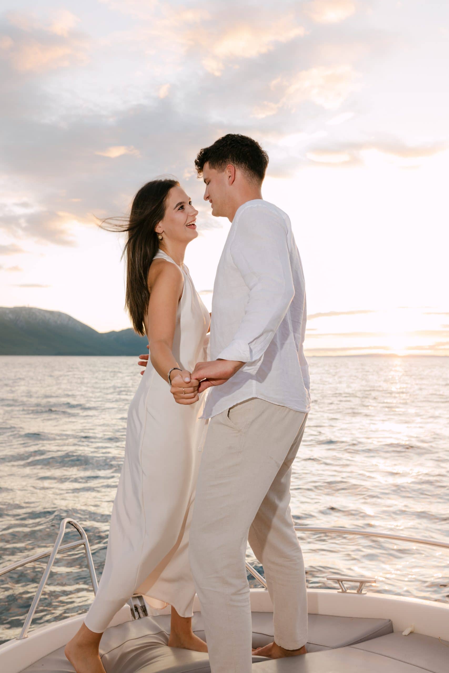 Mladenci se drže za ruke i smiju na brodu tijekom zalaska sunca. Romantična i prozračna fotografija zaruka na moru u Hrvatskoj.